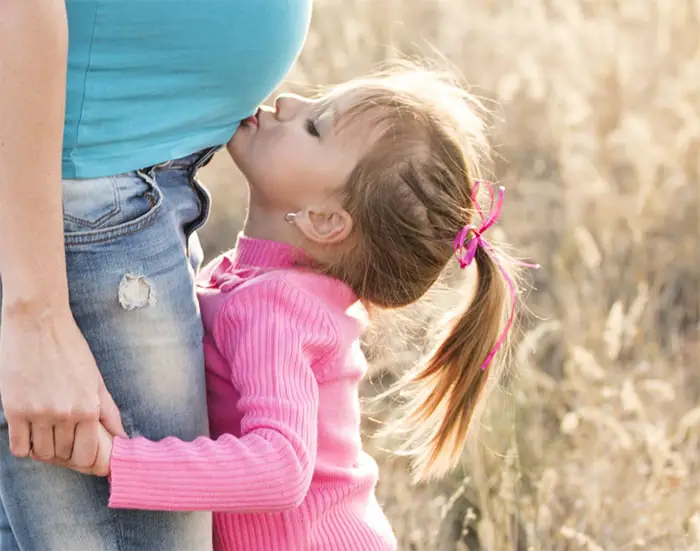 Little girl is kissing the tummy of her pregnant mother and the mother is wearing the best maternity jeans