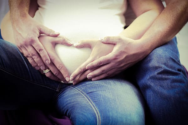 Pregnant women and her husband is touching her tummy while doing a heart shape using their fingers and the pregnant woman is wearing the best maternity jeans